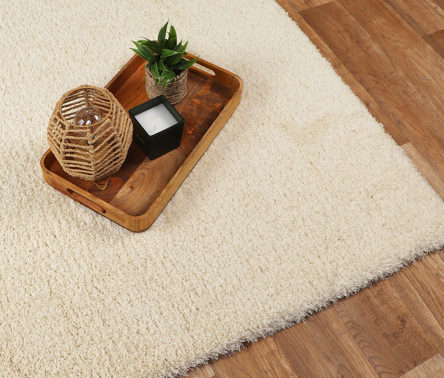 Ein creme farbener Teppich »Pleasure« liegt auf einem Holzboden. Auf dem Teppich steht ein Holztablett mit einer Kerze, einer Pflanze und einer Lampe.