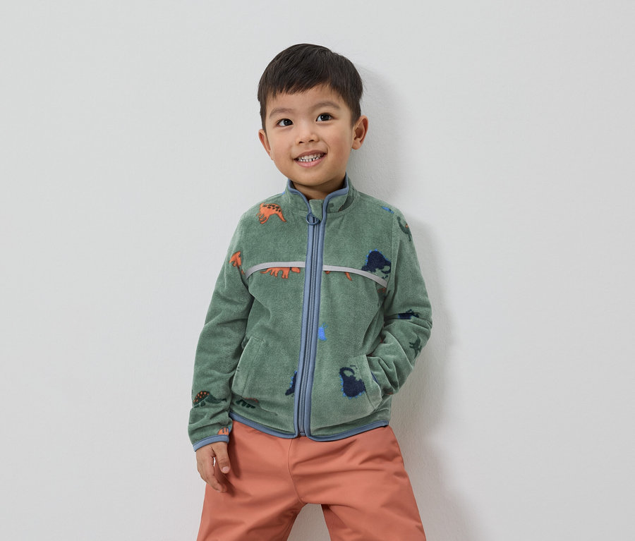 Ein lächelnder Junge mit braunen Haaren trägt eine grüne Fleecejacke mit Dinosaurier-Motiv und orangefarbene Hosen.