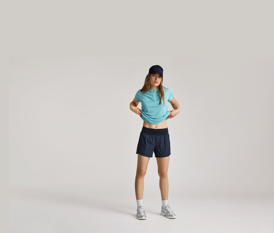 Frau mit marineblauer Kappe, hellblauem Shirt und Shorts, die ihr Shirt anhebt, mit weißen Socken und silbernen Turnschuhen.
