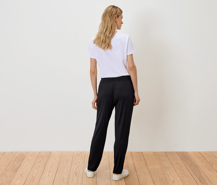 Rückansicht einer Frau mit blonden, gewellten Haaren, die ein weißes T-Shirt, eine schwarze Hose und weiße Turnschuhe trägt.