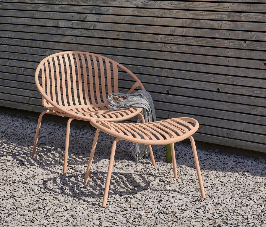 Ein lachsfarbener Stuhl und eine Fußbank stehen auf Kies vor einer Holzwand. Auf dem Stuhl liegen eine Sonnenbrille und eine graue Decke.