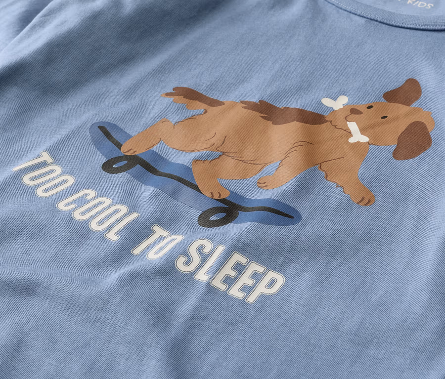 Hellblaues T-Shirt mit einem braunen Hund, der auf einem blauen Skateboard liegt und einen Knochen im Maul hält. Darunter steht der weiße Text "TOO COOL TO SLEEP".