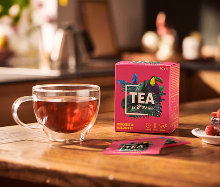 Auf einem Holztisch stehen eine Glastasse mit Früchtetee, eine Packung TEA by Tchibo Früchtetee Waldbeere und ein Teller mit Dessert.