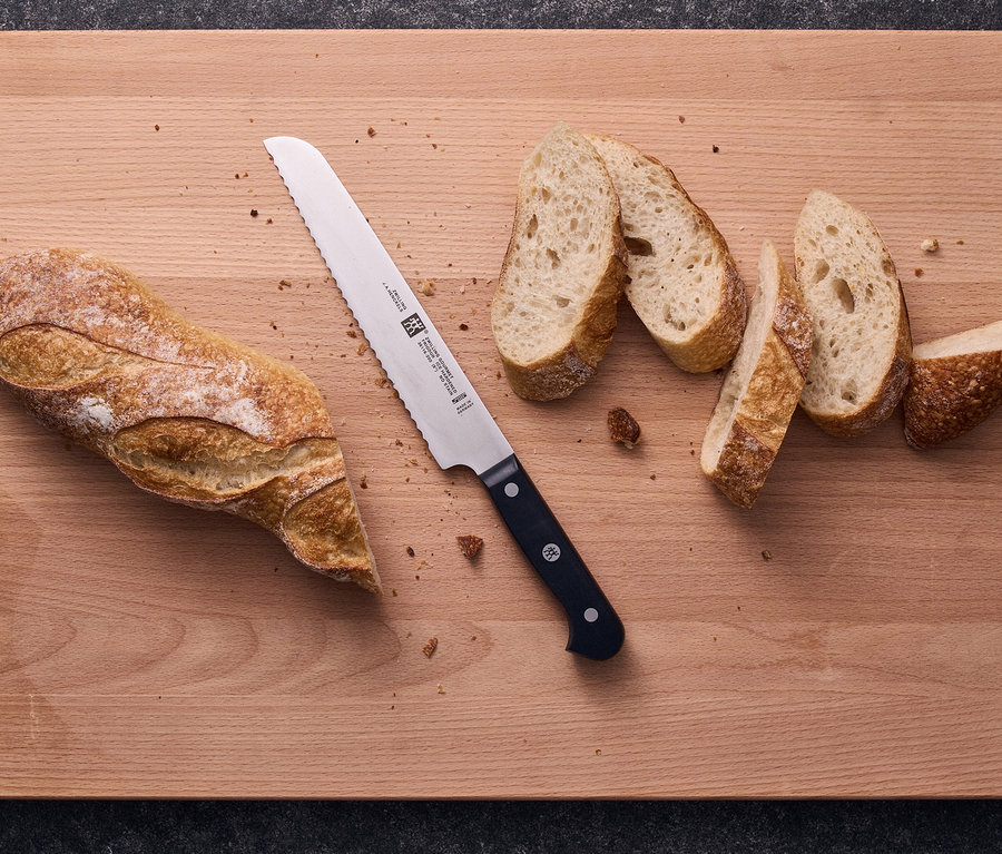 Ein teilweise aufgeschnittenes Baguette und ein Brotmesser liegen auf einem Holzschneidebrett, mit einigen Brotkrümeln verstreut.