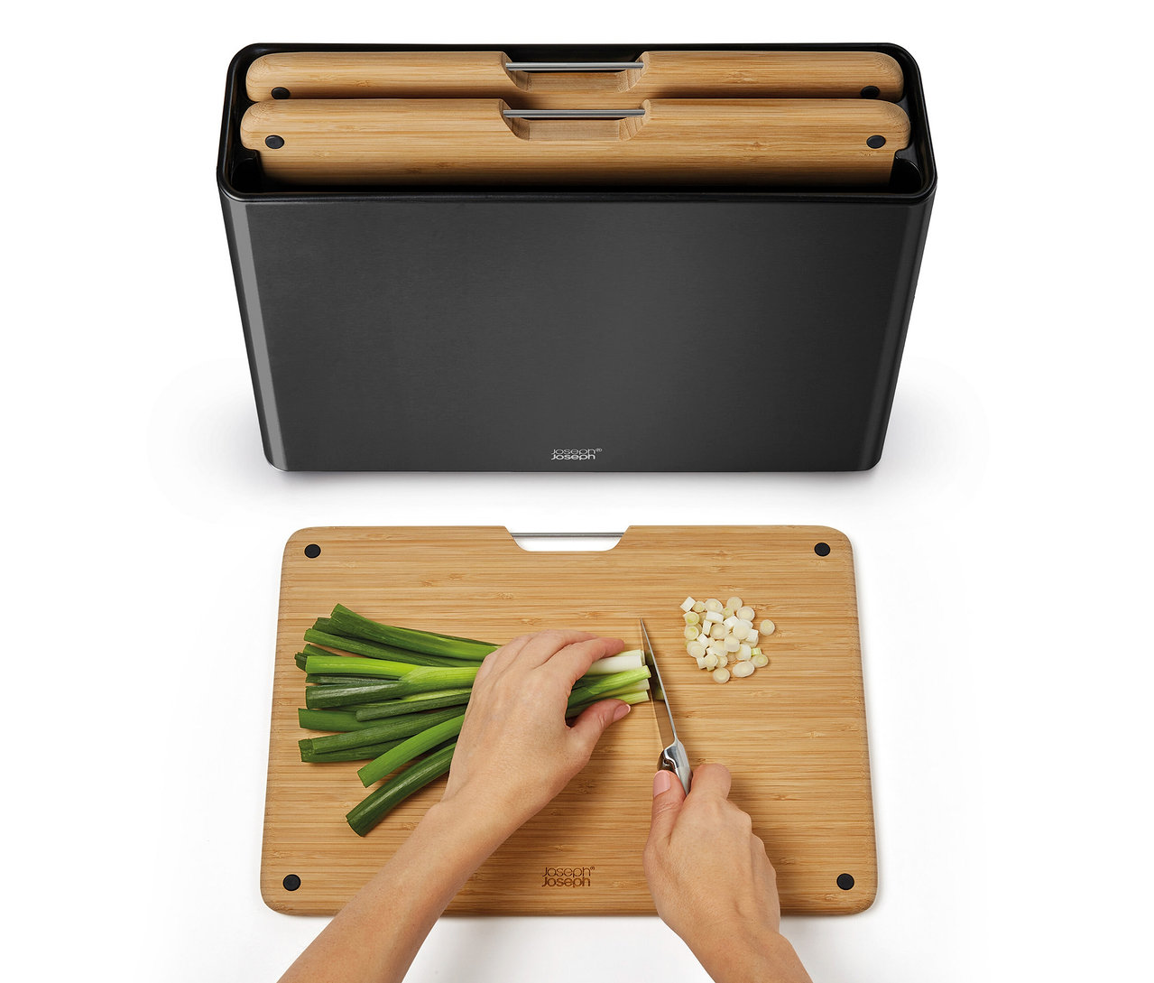 Hände schneiden Frühlingszwiebeln auf einem Joseph Joseph Folio™ Steel Bamboo Schneidebrett, darüber ein schwarzer Halter mit weiteren Brettern.