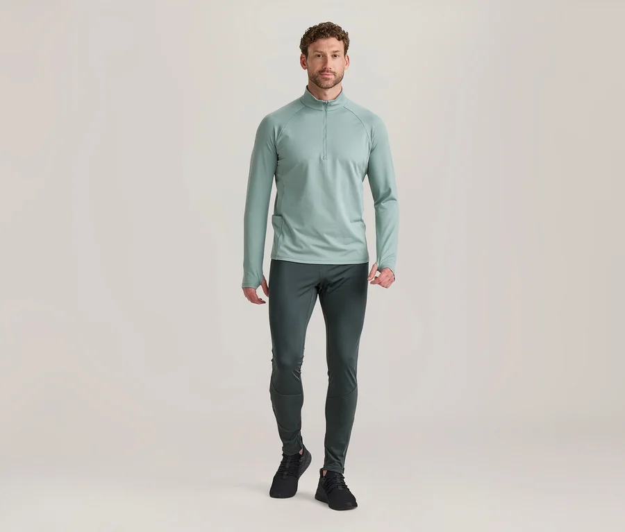 Mann steht in hellgrünem Langarm-Half-Zip-Oberteil, dunkelgrauer Sporthose und schwarzen Turnschuhen.