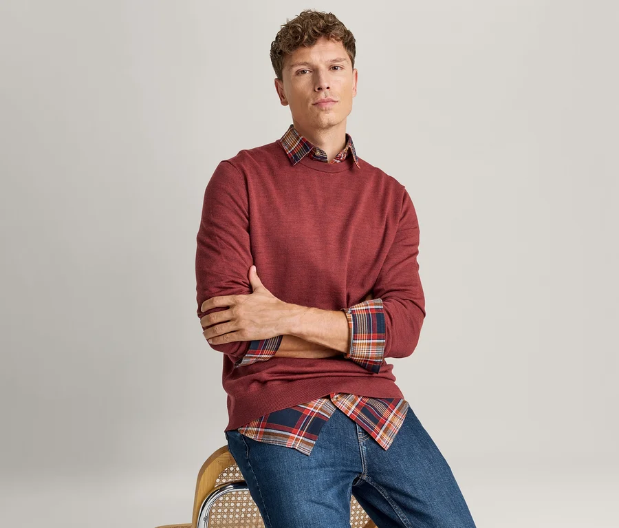 Mann sitzt mit verschränkten Armen, trägt einen roten Langarmpullover über einem rot-blau-orange karierten Hemd und blaue Jeans.