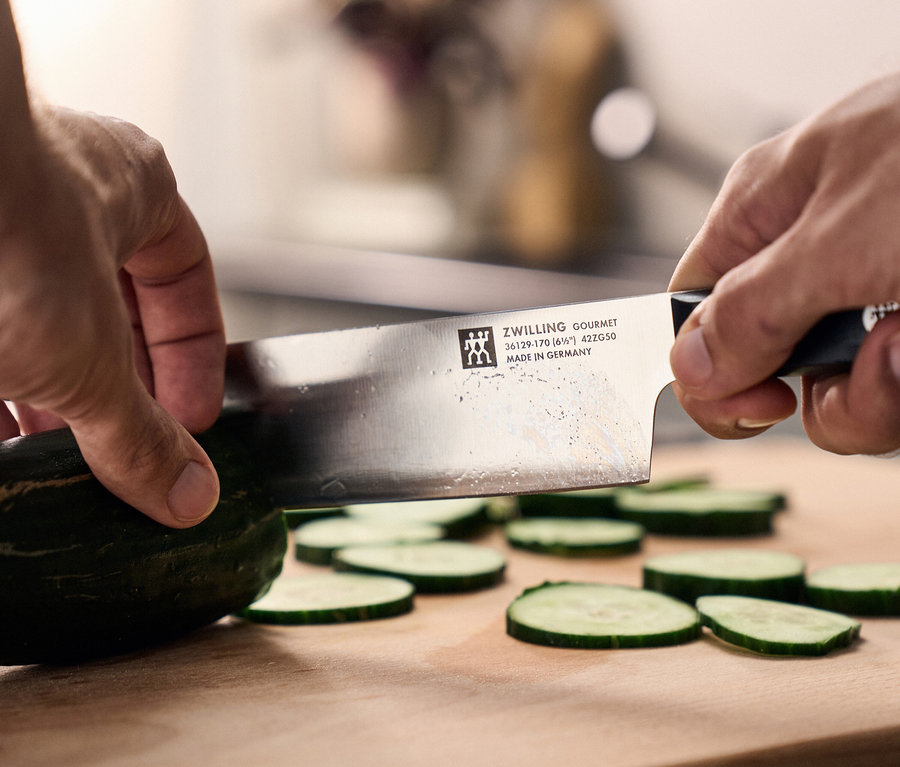 Hände schneiden eine grüne Gurke mit einem ZWILLING GOURMET Messer, hergestellt in Deutschland, auf einem Holzschneidebrett, daneben liegen bereits mehrere Gurkenscheiben.