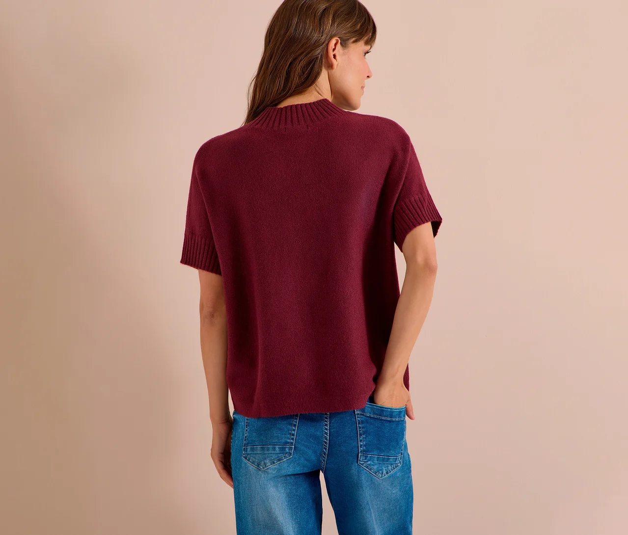 Frau von hinten, trägt einen bordeauxroten CECIL Strickpullover mit kurzen Ärmeln und blaue Jeans. Ihre rechte Hand steckt in der Hosentasche.