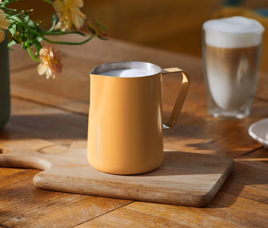 Ein gelber Milchschaumkrug mit aufgeschäumter Milch steht auf einem Holzschneidebrett auf einem Holztisch. Im Hintergrund sind ein Glas Latte Macchiato, eine Vase mit hellen Blumen und ein Teller mit Keksen zu sehen.