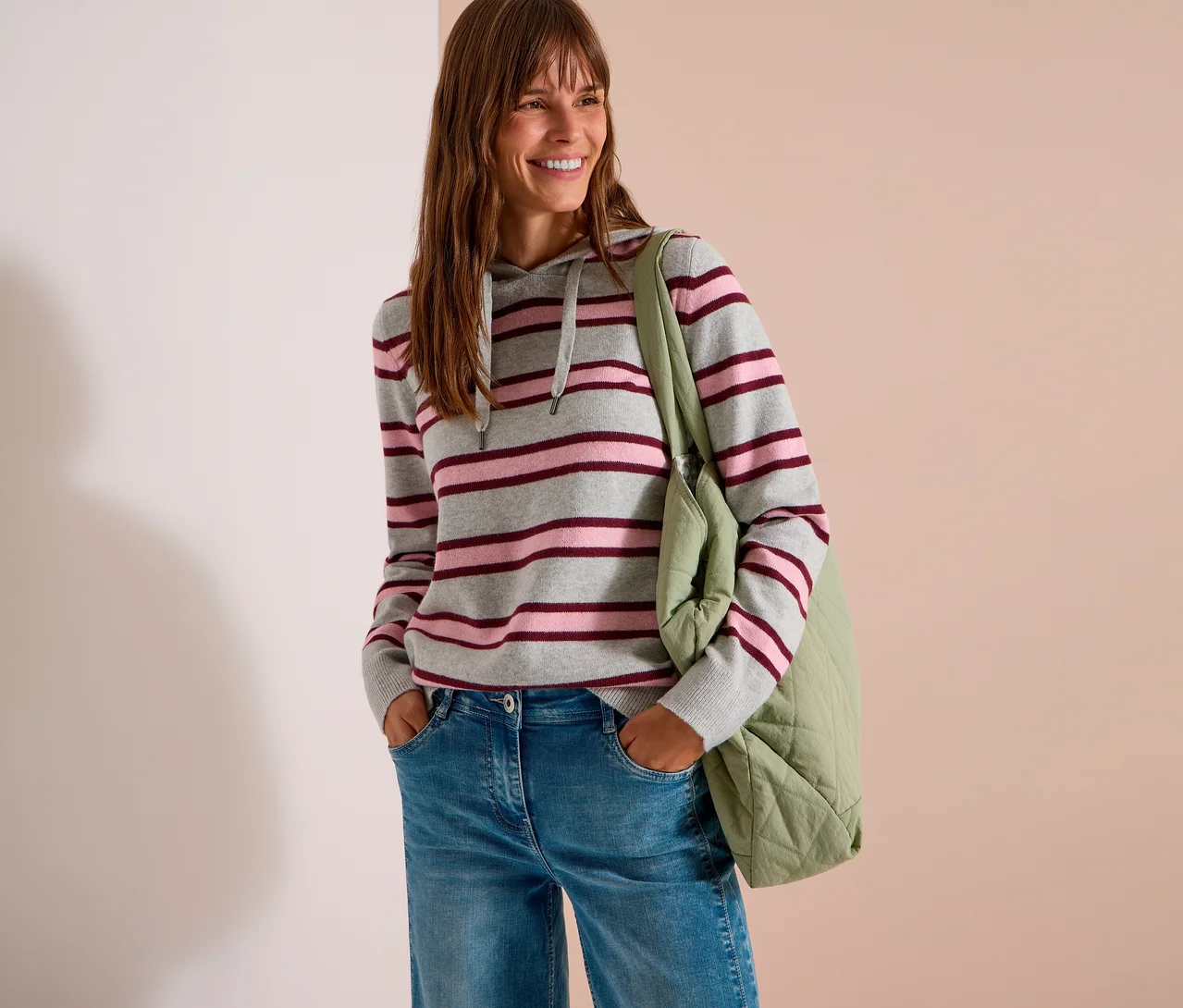 Eine lächelnde Frau mit langen braunen Haaren trägt einen grau, rosa und bordeauxrot gestreiften CECIL Strick-Hoodie, blaue Jeans und eine hellgrüne gesteppte Tragetasche. Ihre Hände stecken in den Hosentaschen.