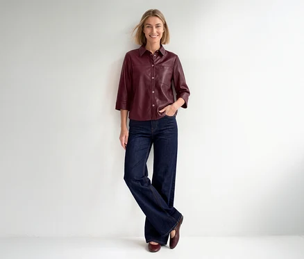 Eine lächelnde Frau steht vor einer weißen Wand und trägt eine bordeauxrote Street One 3/4-Arm-Bluse, weite Jeans und flache Schuhe.