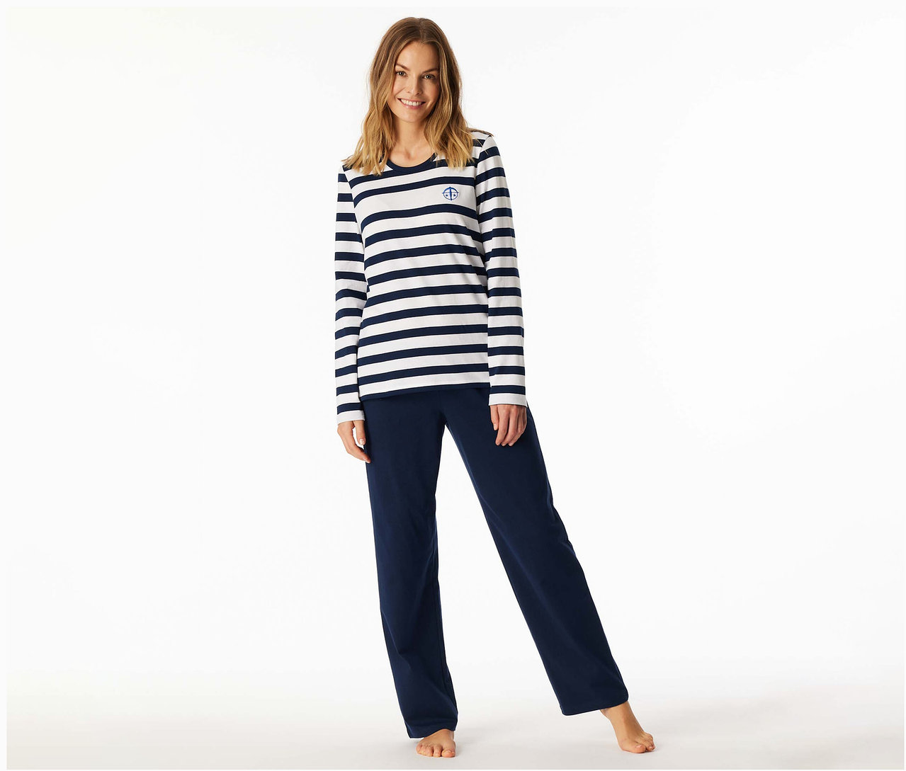Lächelnde Frau trägt ein SCHIESSER Pyjama-Set mit einem weiß-marineblau gestreiften Langarmshirt und einer einfarbigen marineblauen Hose, barfuß stehend.
