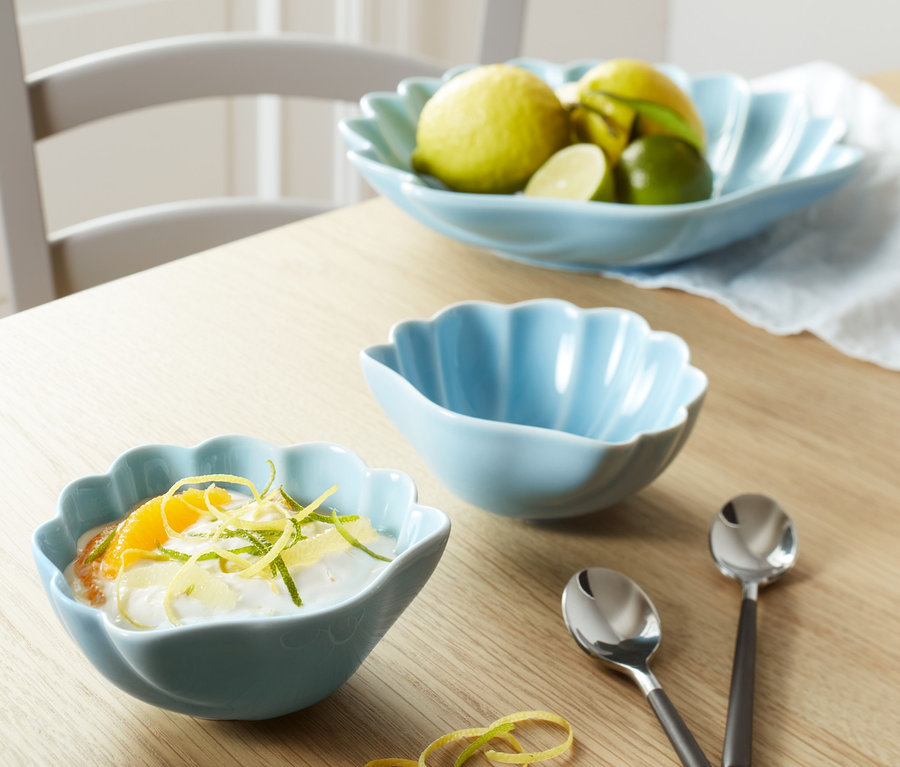 Auf einem Tisch stehen zwei blaue Schalen und Löffel. Eine Schale ist mit Dessert gefüllt, die andere ist leer. Im Hintergrund befindet sich eine größere Schale mit Obst.