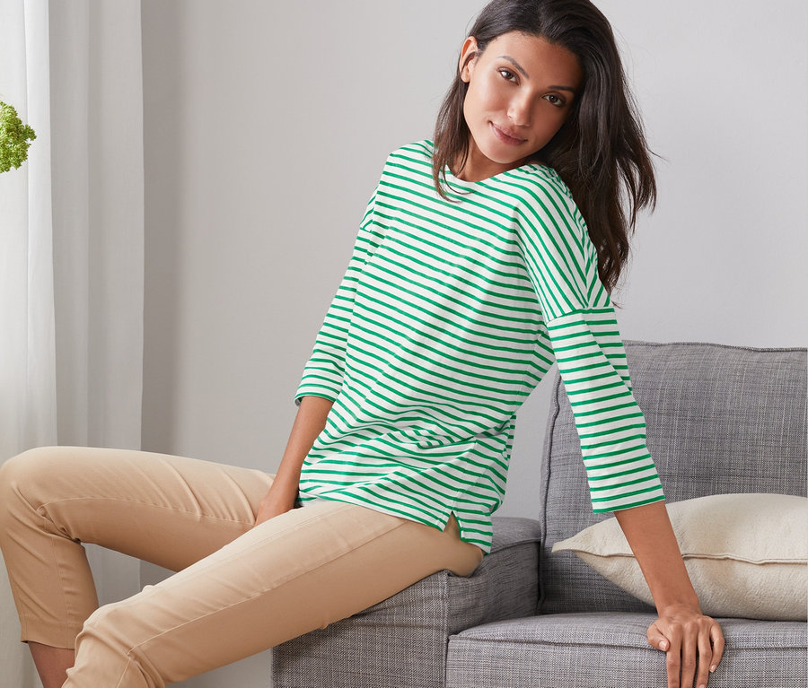 Eine Frau sitzt auf einem grauen Sofa und trägt ein gestreiftes Shirt mit 3/4-Arm.