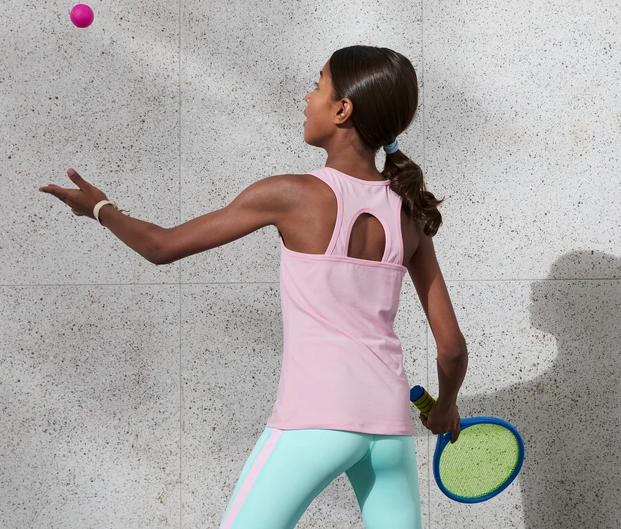 Mädchen in 2 Sport-Tops aus recyceltem Material spielt mit Schläger und Ball.