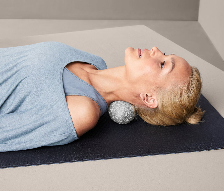 Frau liegt auf einer Matte, mit einem Duo-Faszienmassageball unter ihrem Nacken.