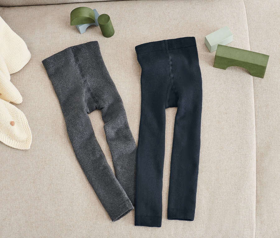 Zwei Paar Thermoleggings in Blau und Grau liegen nebeneinander auf einem beigen Sofa. Auf dem Sofa liegen auch eine beige Strickdecke und grüne Spielsachen.