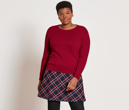 Eine Frau steht und trägt einen roten Zopfstrickpullover, einen karierten Jacquard-Rock, 2 Paar blickdichte Strumpfhosen in Schwarz und Lederboots.