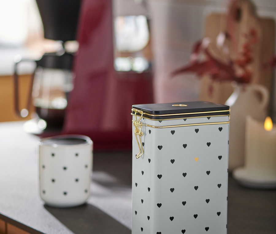 Eine Kaffeedose mit Herzchen steht neben einer Tasse und einer Kaffeemaschine auf einer Küchentheke.