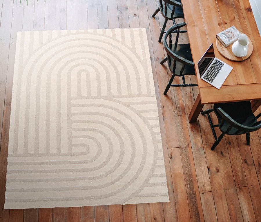 Draufsicht eines beigen Teppichs »Cosmo« mit geometrischem Bogenmuster auf einem Holzboden, daneben ein Holztisch mit Laptop und Stühlen.