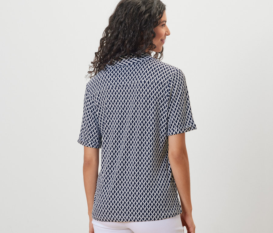 Rückansicht einer Frau mit dunklem, gewelltem Haar, die ein bedrucktes Blusenshirt mit Knopfleiste und eine weiße Hose trägt.