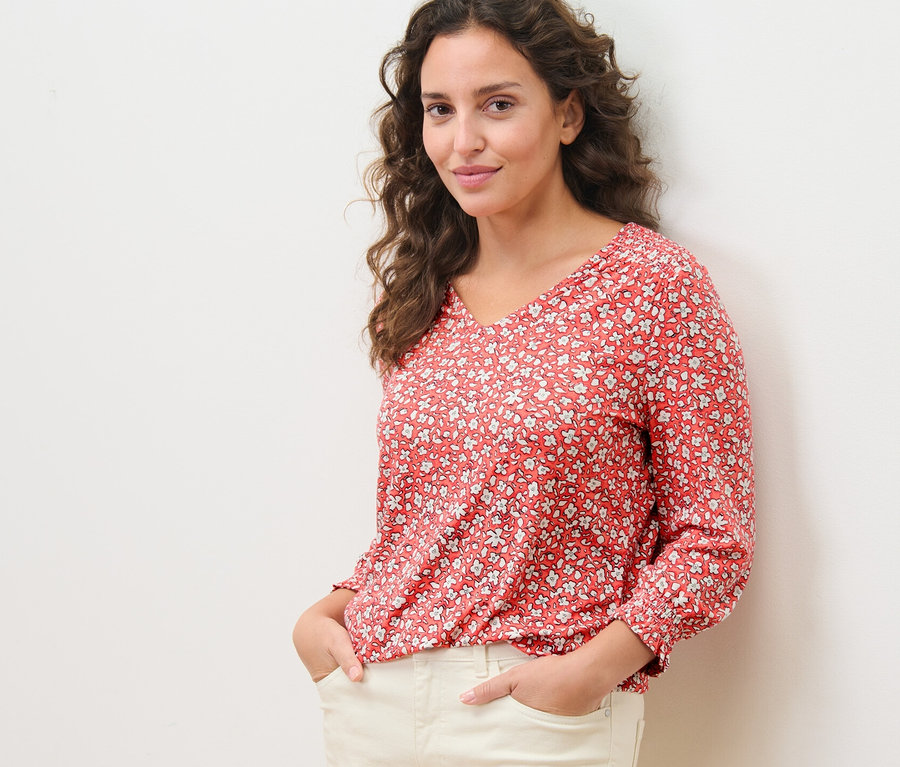 Frau posiert in einem korallenfarbenen, bedruckten Blusenshirt und heller Hose.