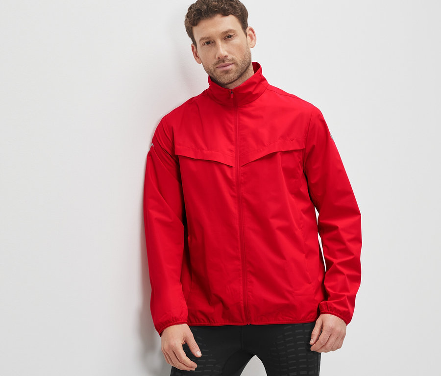 Mann lehnt an Wand, trägt rote Windprotection-Funktionsjacke.
