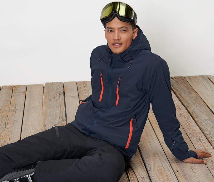 Mann sitzt auf Holzboden und trägt eine High-Tech-Skijacke und Skibrille.