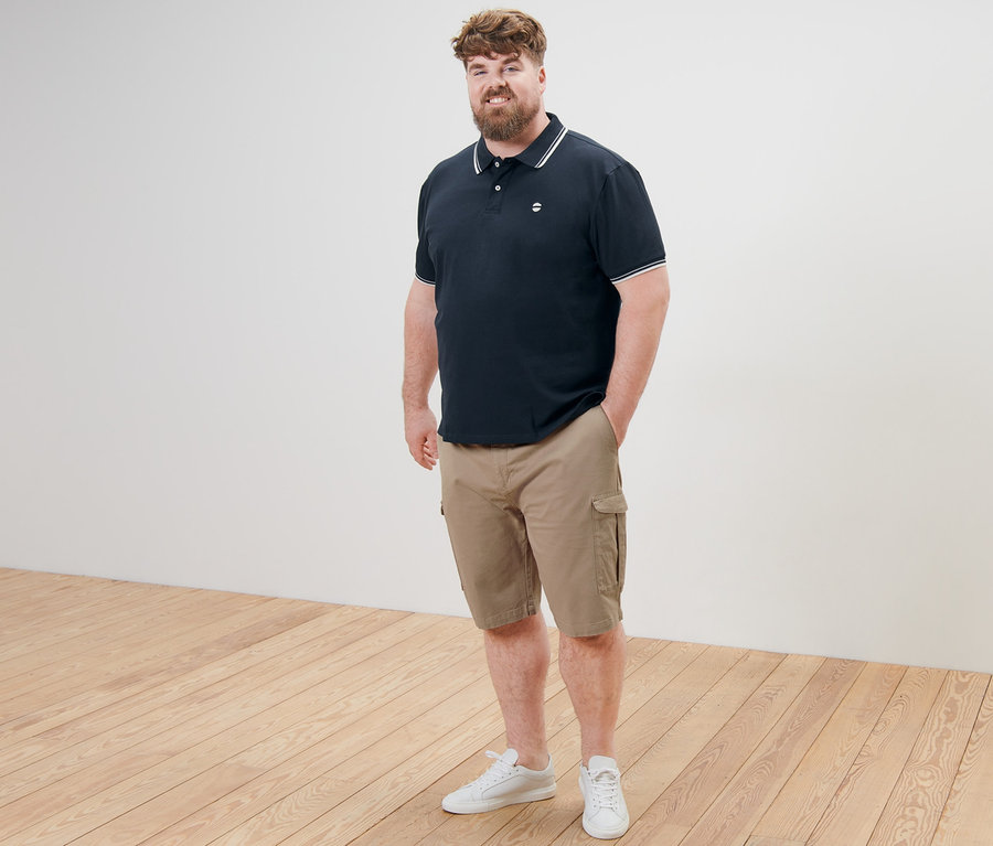 Mann in marineblauem Piqué-Poloshirt und beigen Cargo-Shorts steht mit einer Hand in der Hosentasche.