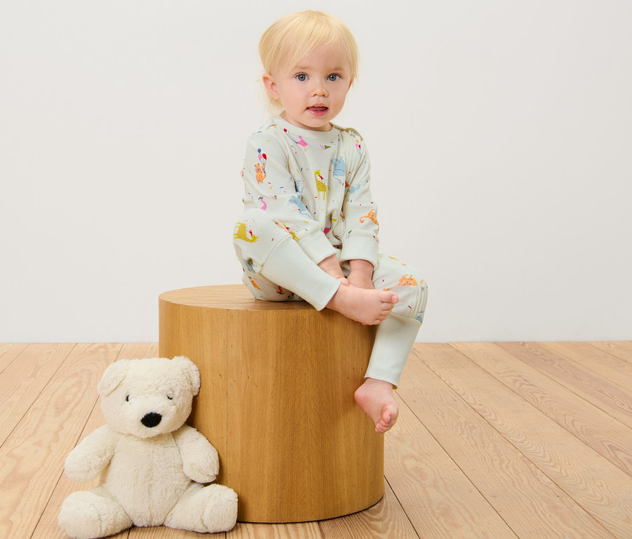 Ein Kind sitzt auf einem Holzhocker und trägt einen 3 Baby-Pyjama mit Tiermotiv, daneben liegt ein Teddybär.
