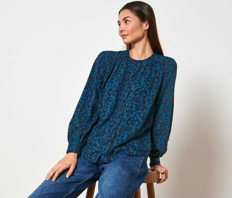 Frau sitzt auf einem Hocker und trägt eine Bluse mit Alloverprint und Jeans.