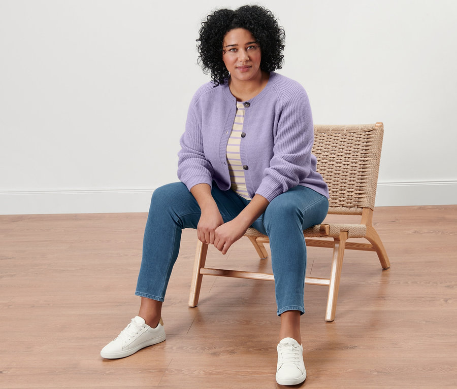 Frau sitzt auf einem Korbstuhl und trägt ein beige-fliederfarben gestreiftes Shirt mit 3/4-Arm, einen Strickcardigan, Jeggings und Ledersneaker.