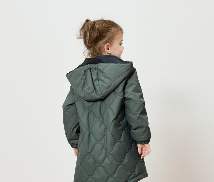 Von hinten ist ein kleines Mädchen mit zu einem Pferdeschwanz zusammengebundenen Haaren zu sehen, das eine grüne Steppjacke mit Kapuze trägt.