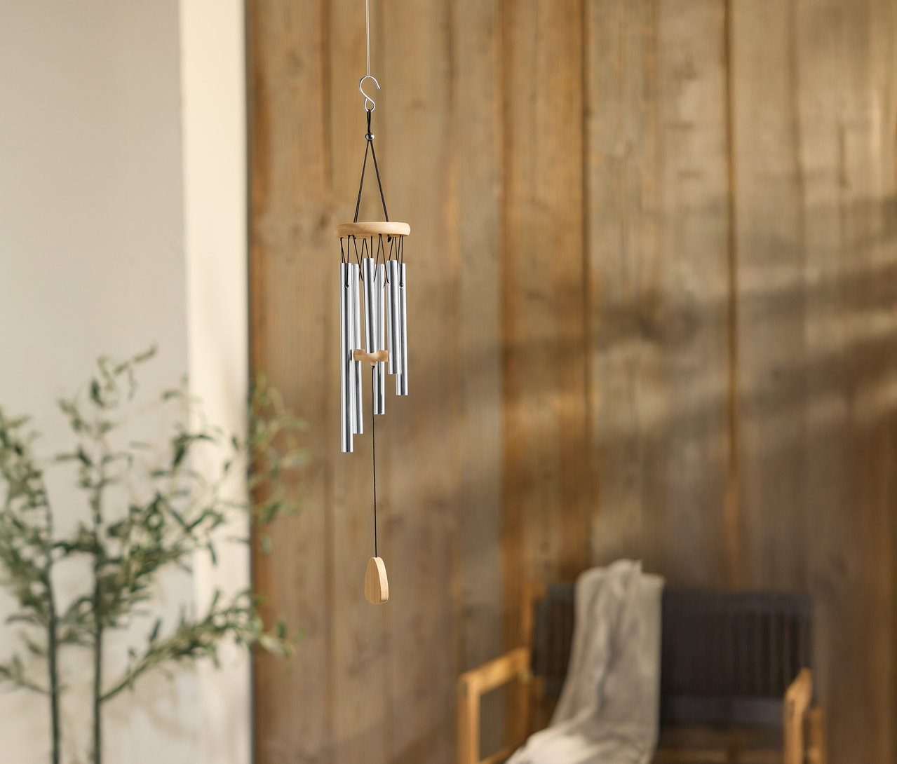 Ein Windspiel mit silbernen Rohren hängt vor einer Holzwand, neben einer Pflanze und einem Stuhl.