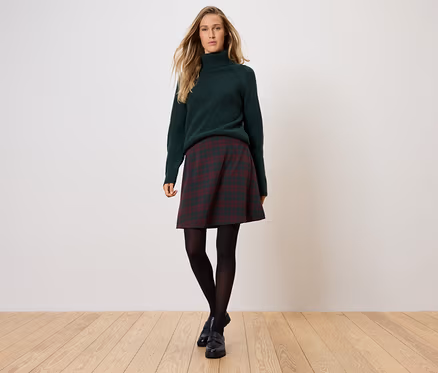 Frau steht und trägt einen grünen Rollkragenpullover, einen karierten Rock, eine schwarze Strumpfhose und schwarze Schuhe.