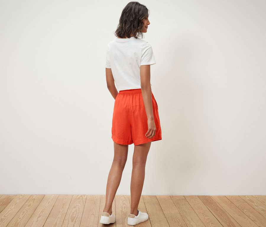 Frau steht mit dem Rücken zur Kamera und trägt orange Leinenshorts und ein weißes T-Shirt.