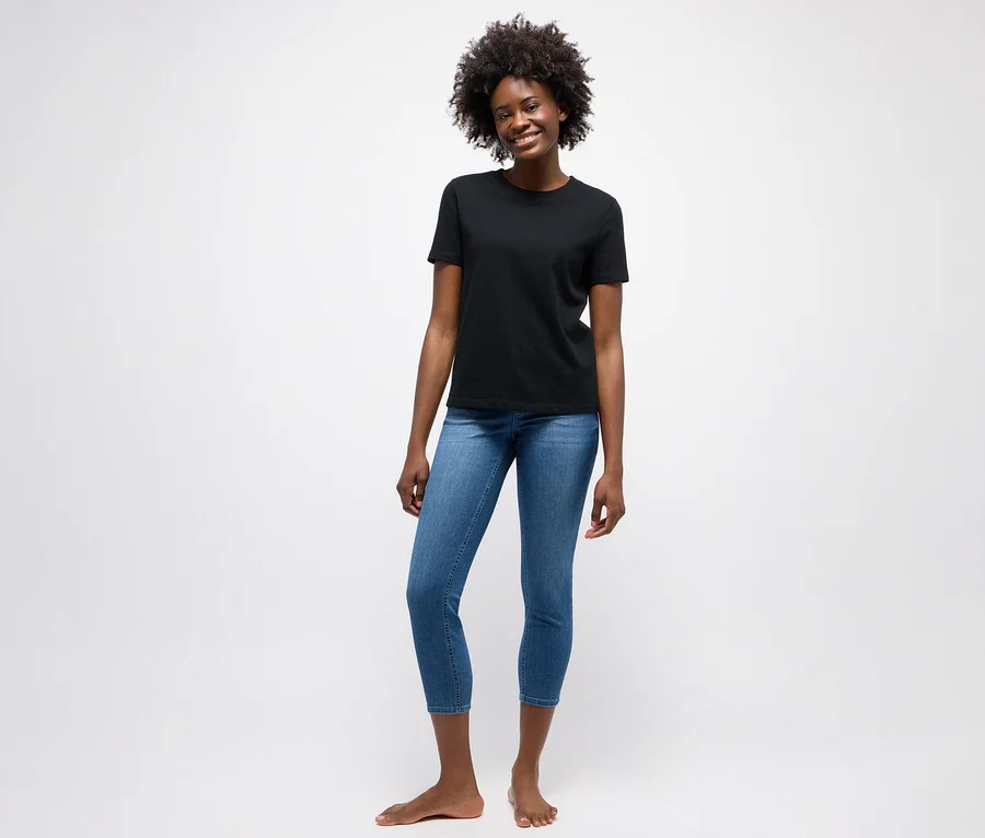 Lächelnde Frau mit Locken trägt ein schwarzes T-Shirt und eine mittelblaue Angels Jeans »Ornella« Slim Fit 7/8, barfuß stehend.