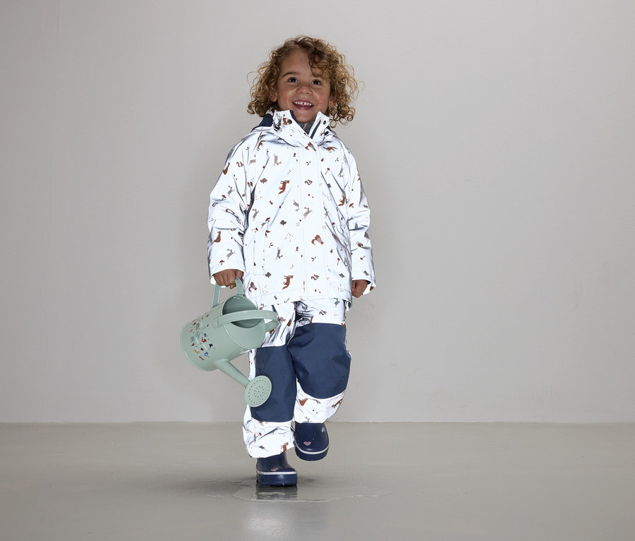 Ein Kind mit lockigem Haar trägt eine Jacke und Hose mit Tiermuster und hält eine grüne Gießkanne.