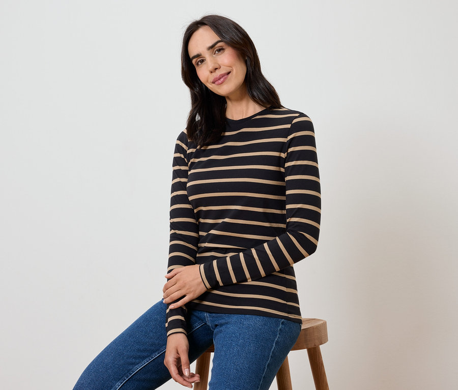 Eine Frau sitzt auf einem Hocker und trägt ein schwarz-beige gestreiftes Langarmshirt und blaue Jeans.