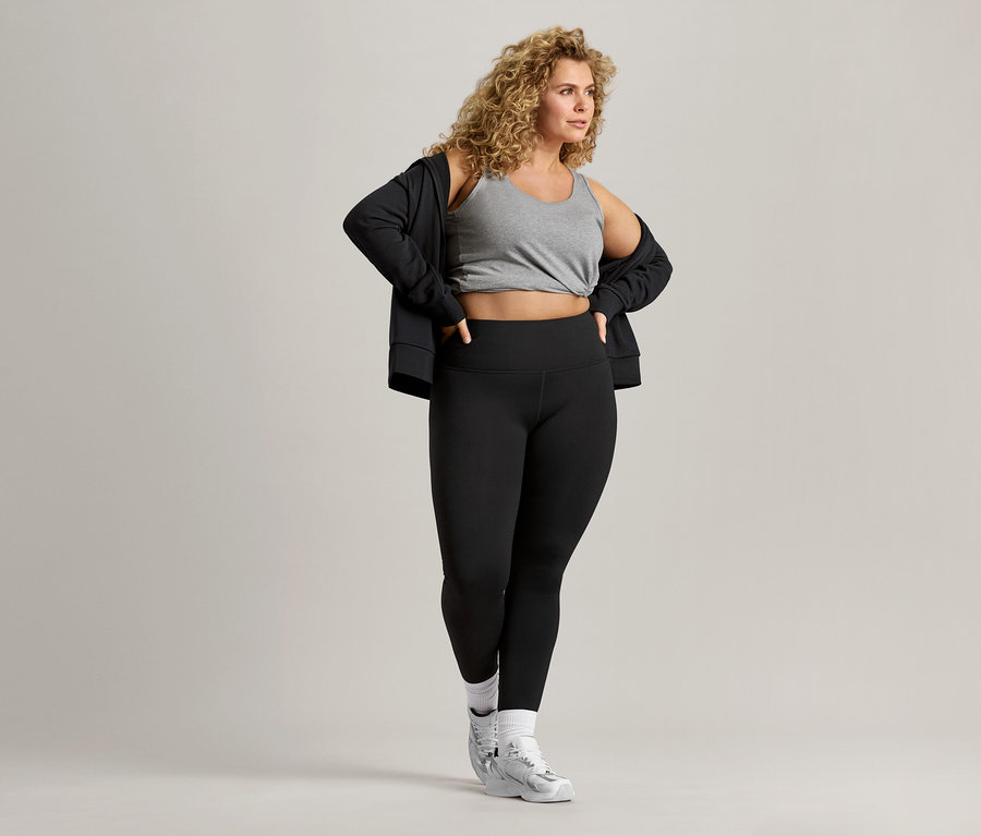 Frau mit lockigem Haar trägt ein graues Tanktop, eine schwarze Jacke und schwarze Leggings.