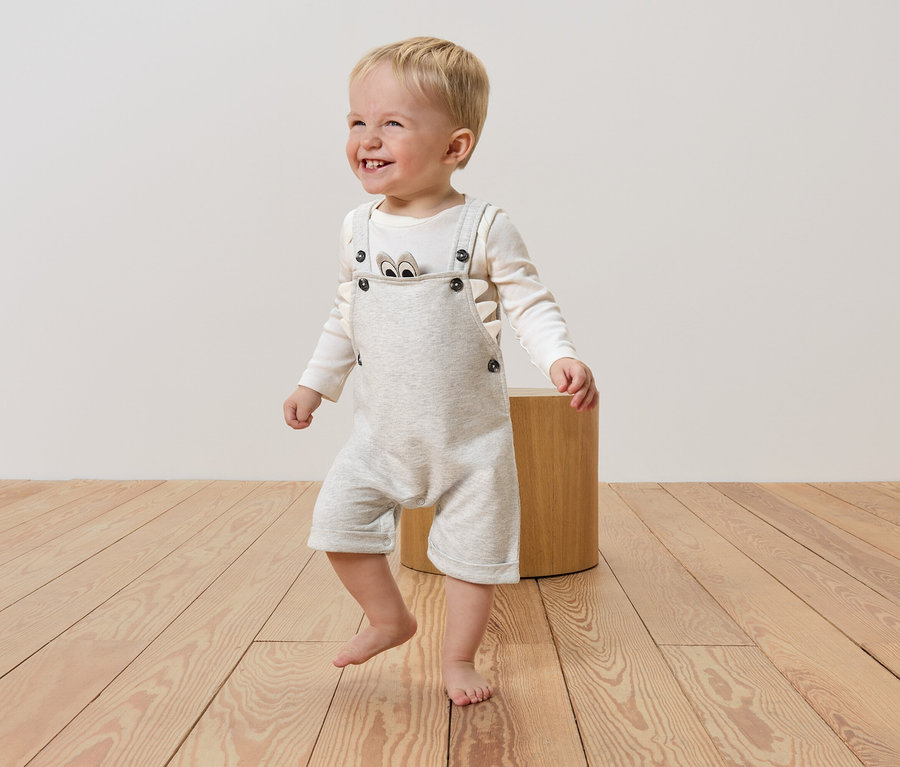 Ein lachender, hellhaariger Junge mit nackten Füßen trägt einen hellgrauen Jumpsuit mit Knöpfen und ein langärmliges Shirt mit einem Bärenaufdruck, während er auf einem Holzboden neben einem Holzhocker steht.