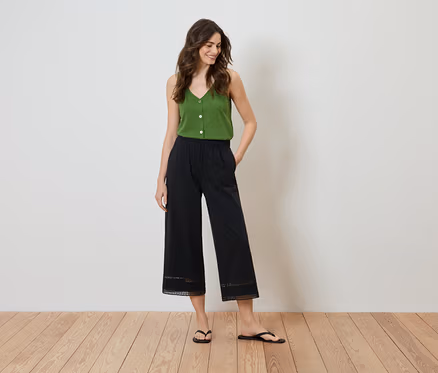 Frau steht mit den Händen in den Taschen, trägt ein grünes Top und eine schwarze Culotte mit Spitzendetail am Saum.