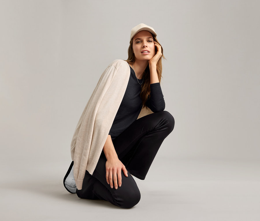 Frau hockt mit schwarzem Outdoor-Funktionsshirt, beiger Strickjacke und heller Kappe.