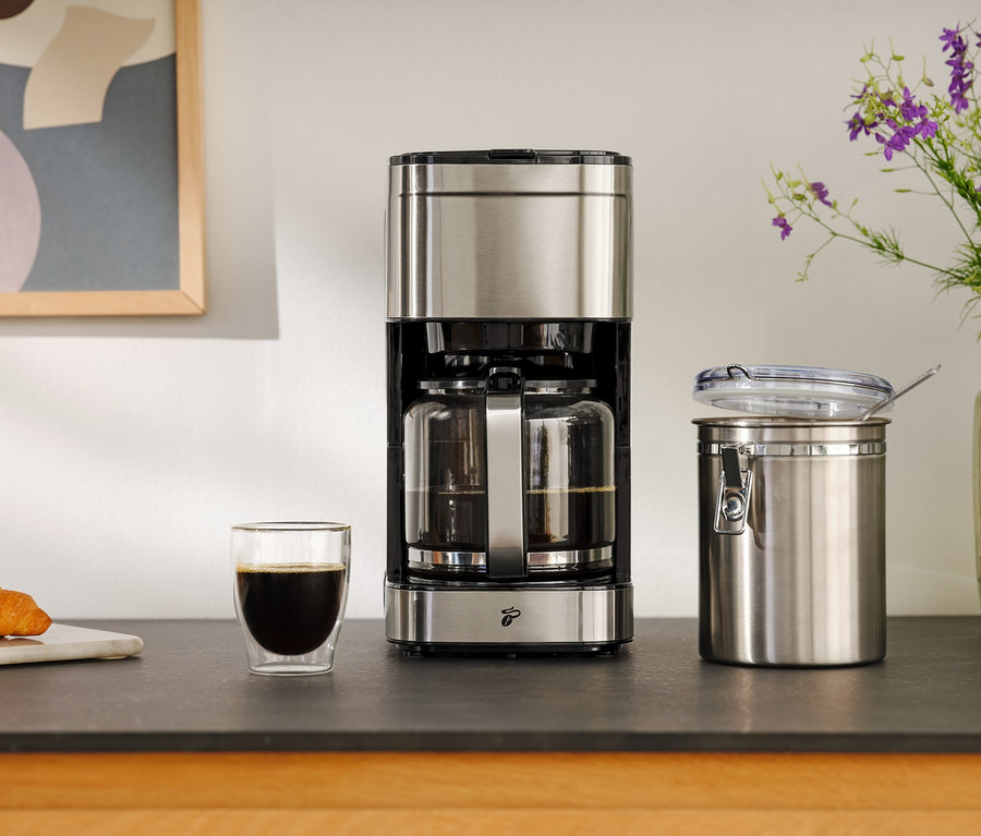 Kaffeemaschine neben einem Glas Kaffee, einem Croissant und einem Vorratsbehälter.