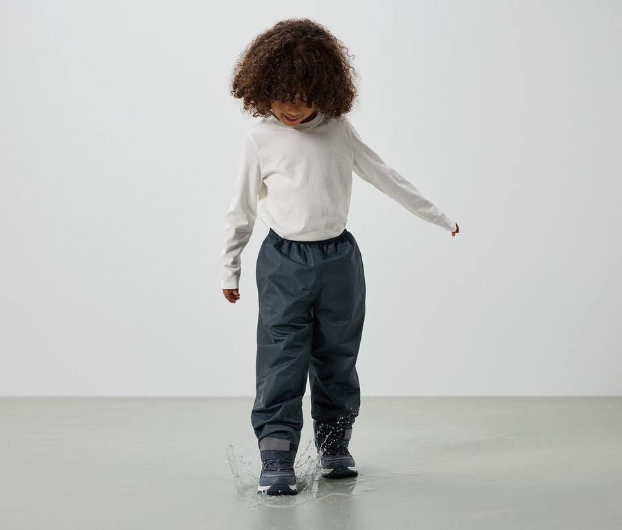 Kind mit lockigem Haar, bekleidet mit einem langärmeligen Oberteil, grauen Regenhosen und Stiefeln, springt in eine Wasserpfütze.
