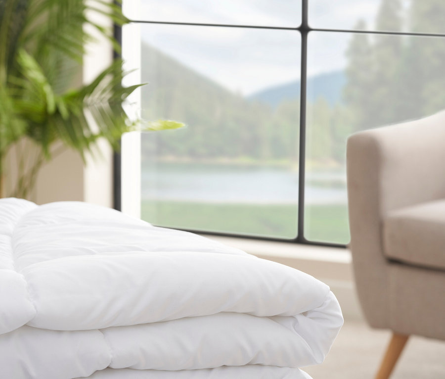 Ein weißer, gesteppter Bettüberwurf liegt auf einem Hocker vor einem Fenster mit Blick auf einen See.