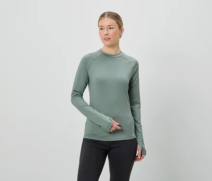Frau mit blonden Haaren trägt ein grünes, langärmliges Oberteil und schwarze Leggings vor einem weißen Hintergrund.
