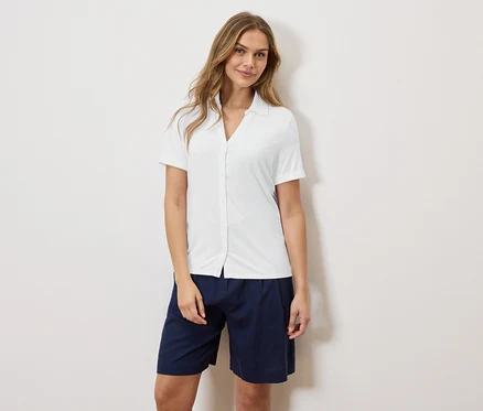 Eine Frau steht vor einer Wand und trägt ein weißes Hemd und blaue Shorts.