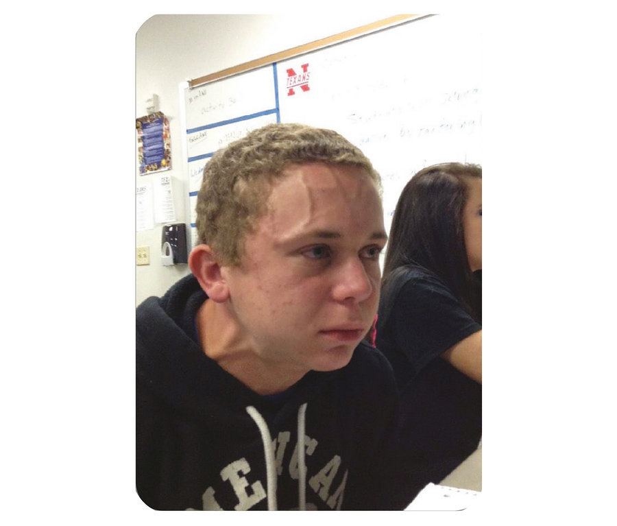 Ein junger Mann mit angespannten Gesichtszügen und hervortretenden Adern auf der Stirn sitzt in einem Klassenzimmer.